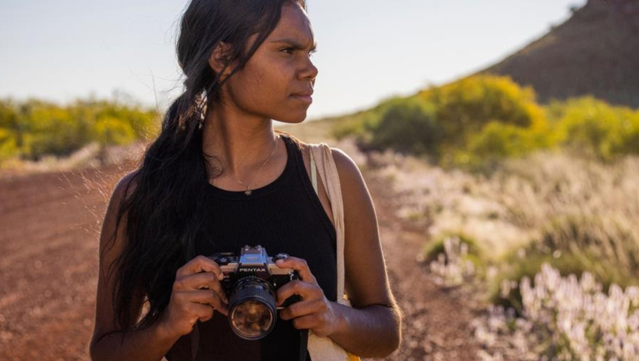 Young Indigenous woman with camera on desert road, a product of Screenwest and West Coast Visions.