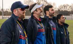 Four men stand beside a football pitch in Ted Lasso