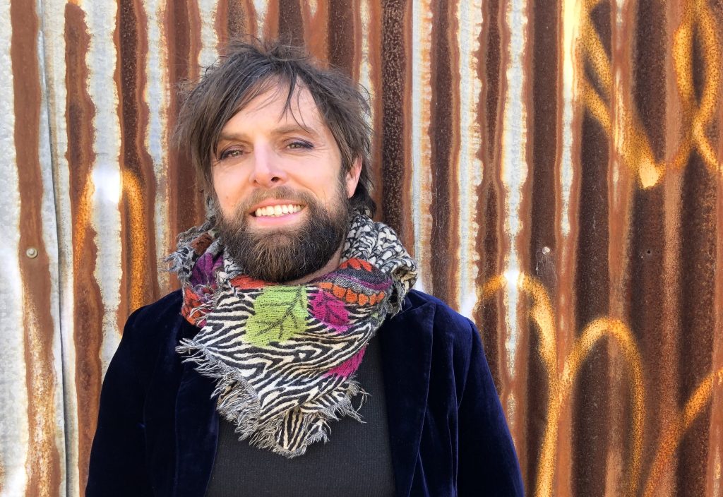 Paul Dalgarno in a scarf stands against a rusty corrugated iron backdrop