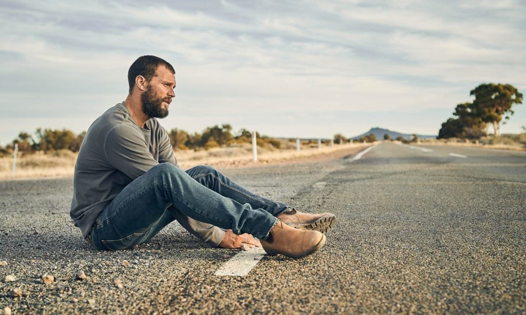 A man in dishevelled clothing sits alone in an empty road