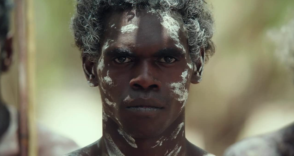 An indigenous man with white paint on his face looks at the camera