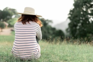 A woman hugs her knees on a hillside, she is seen from behind.