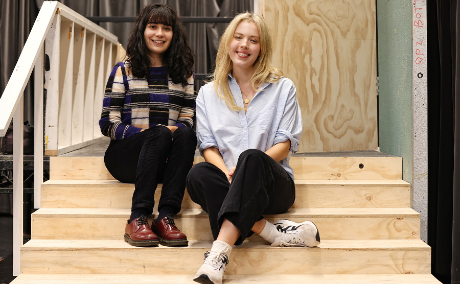 Two women sit on a wooden set smiling at the camera. Both have their hands in their lap.