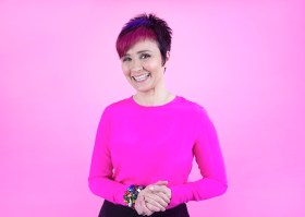 Cal Wilson. Image of woman with short red and black hair, wearing pink jumper and clasping hands.