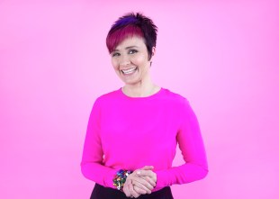 Cal Wilson. Image of woman with short red and black hair, wearing pink jumper and clasping hands.