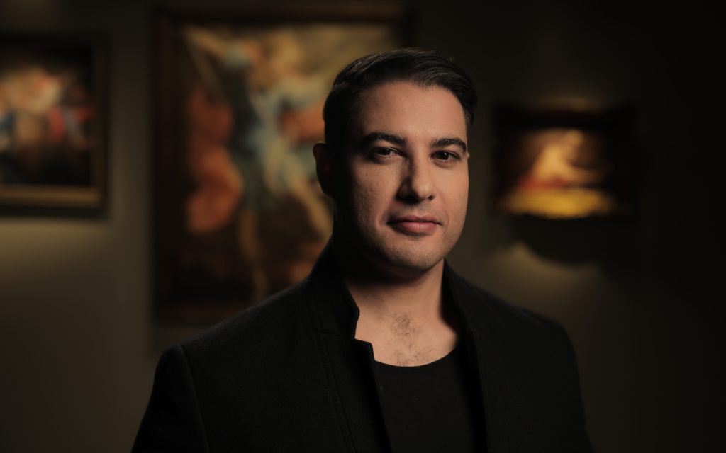 The Mission. Image is of a man in black T-shirt and jacket with short dark hair, looking at the camera, standing in front of a dark backdrop including blurry Old Master paintings.
