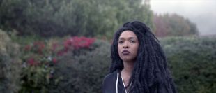 Still from 'The Tuba Thieves'. Image: TOFF. A figure with long curly black hair and brown skin wearing a black top stands in a garden with a moody atmosphere, looking into the distance.