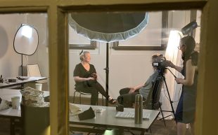 Documentary. Image is a group of women in a small filming studio, with two sitting in chairs facing each other and another behind a film camera.