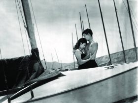Nakahira. image is black and white shot of two young people embracing on a yacht which is moored.