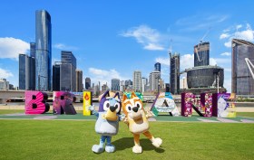 A life-size Bingo and Bluey pose outside the iconic 'Brisbane' sign on the Southbank riverfront.