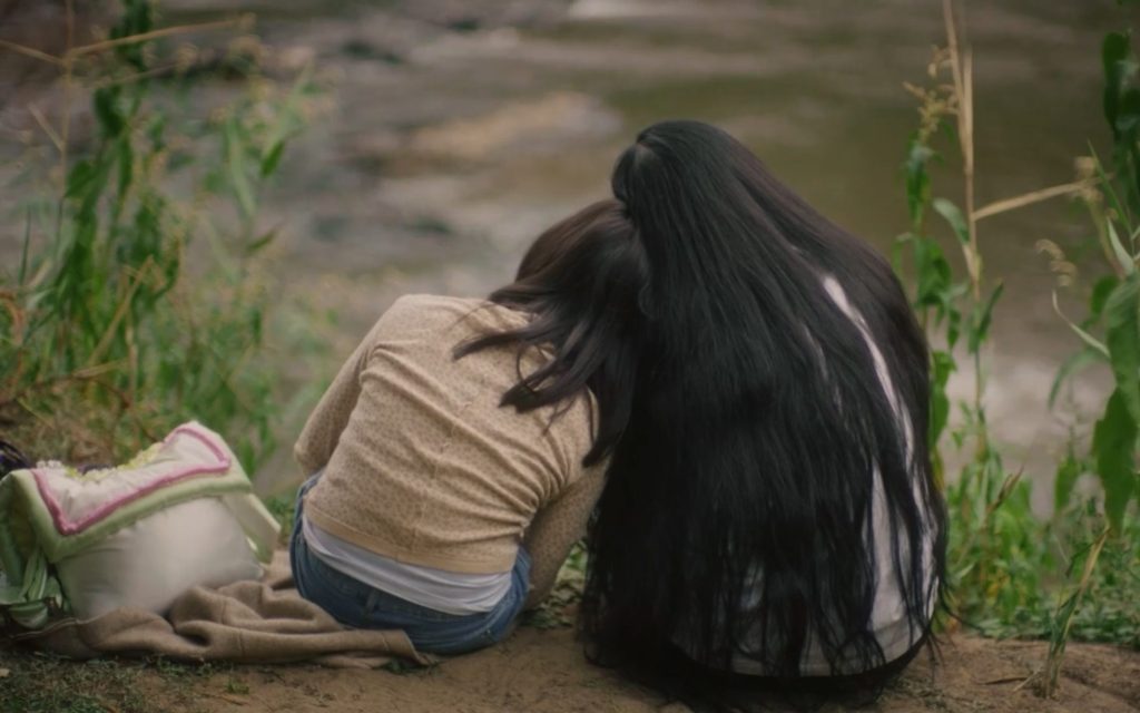 Still from Chi Tran’s film, ‘ The Sun Sets and Once Again the Earth is Upright’. Image: Courtesy the artists. Two women with dark long hair sitting near a river bank with their backs facing the viewer. They are leaning on each other.