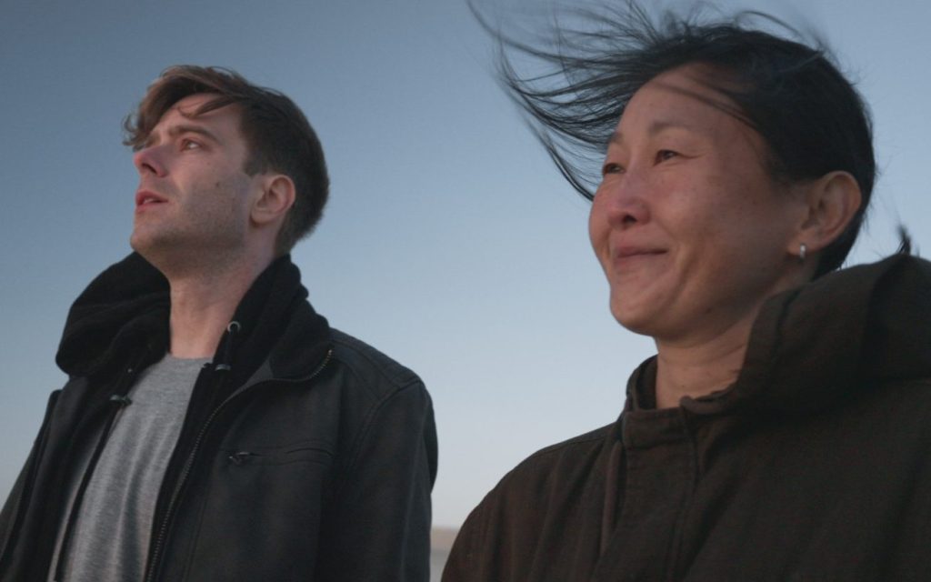 Still from short film 'Jia'. Image: Supplied. A white man stands on the left staring off into the distance, while an Asian woman stands beside him, smiling with hair flowing in the wind. They are both seen from the chest up and wearing black jackets.