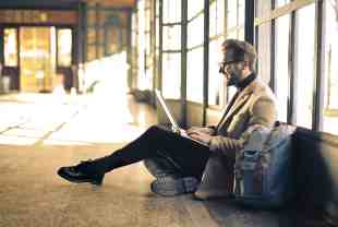 Young man in spectacles wearing turtleneck and blazer sits on floor balancing a laptop on knees, with a backpack at his side.