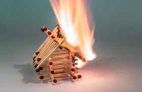 small house made of matches is burning on one side.