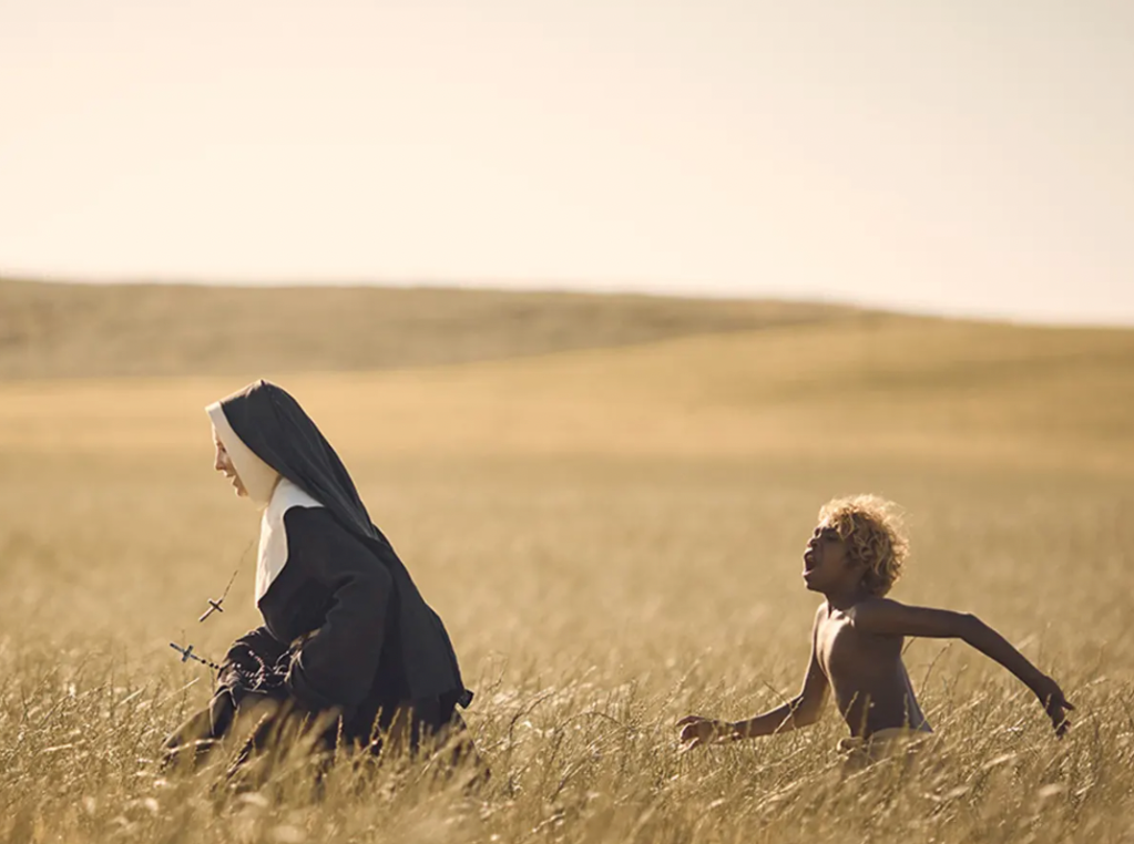 A catholic nun and a young Australian Indigenous boy walk through a field of dry, yellow grass.