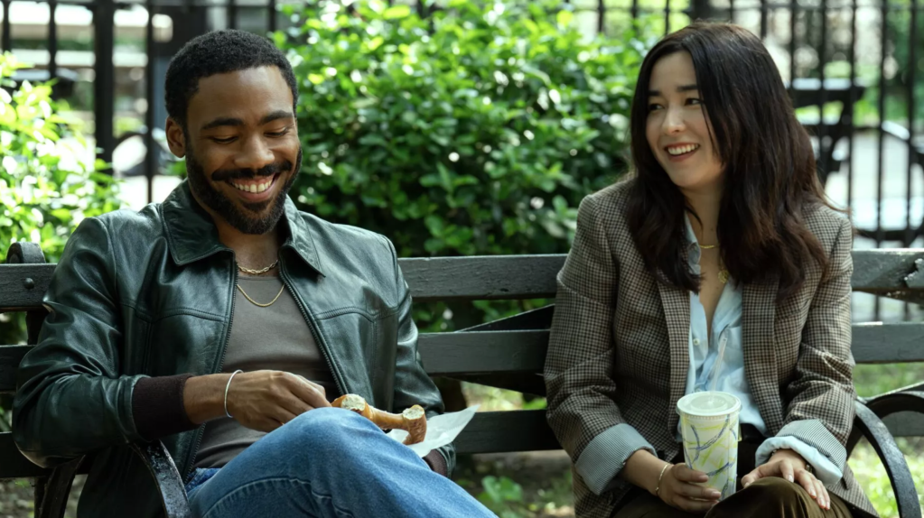 Donald Glover as Mr Smith (wearing a leather jacket) and Maya Erskine as Mrs Smith (wearing a tweed jacket) sit on a park bench in New York.