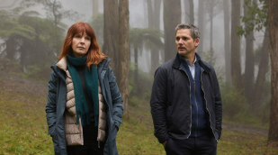 A woman with red hair and a man with grey hair stand in a dense, foggy rainforest.