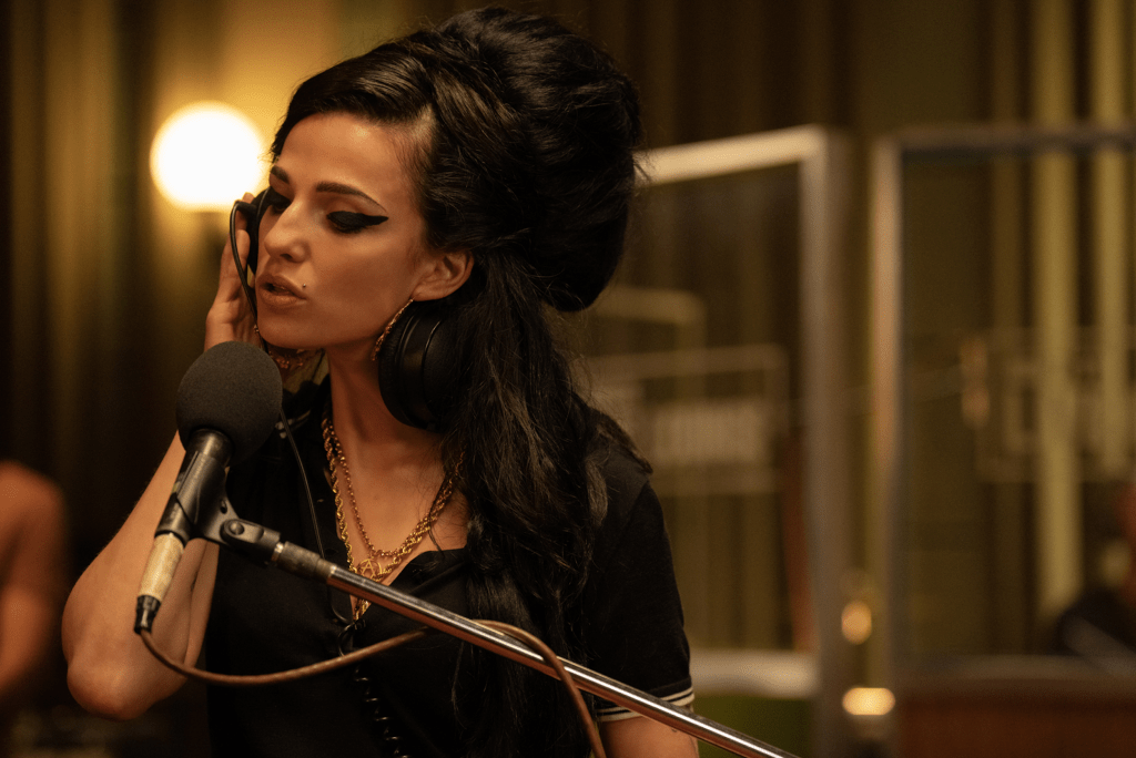 Marisa Abela as Amy Winehouse, a woman with black hair and heavy makeup holding headphones to her ear and singing into a studio microphone.