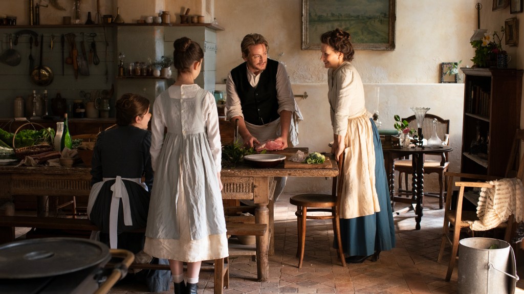 Film still from The Taste of Things. Three women and one man stand around a table in a French provincial kitchen.