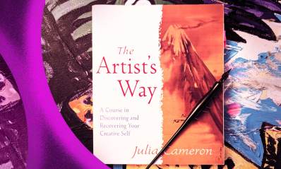 Book cover of The Artist's Way by Julia Cameron, photographed against a background of an oil painting with a calligraphy pen resting on top.