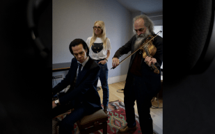 Nick Cave at the piano, Sam Taylor Johnson standing and Warren Ellis playing violin in a studio.