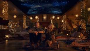 A man and woman sit on a rug on the floor of a house filled with sparkling lights and a ceiling that looks like the night sky in a publicity still for The Big Door Prize Season 2 on Apple TV+.