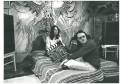 David Tiley. Image is a black and white shot of two long haired white chaps on a bed. One in the bed looks like Jesus, the other is holding a clapperboard. On the wall behind the bed is a huge abstract mural.