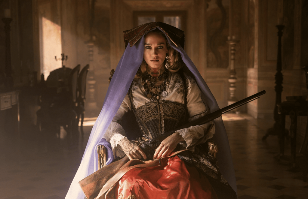 A woman in a red skirt and violet vale with a shotgun on her lap looks directly into the camera in a publicity still for the show Brigands: The Quest for Gold, on Netflix.