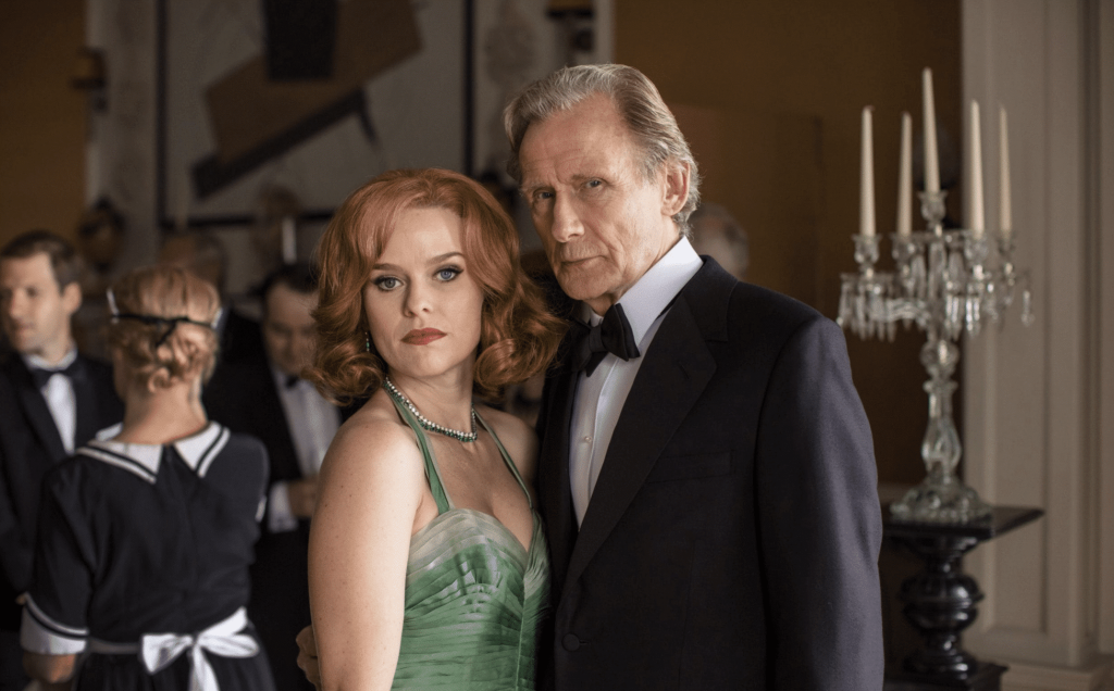 A woman in a green dress and man in a tuxedo look at the camera at a fancy event in a publicity still for Ordeal By Innocence, on BritBox.