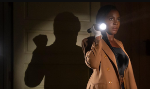 A woman in a brown coat shines a torch in a darkened room in a publicity still for Them: The Scare Season 2 on Prime Video.
