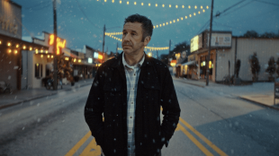 A man walks down the middle of a quiet street in the evening with his hands in his pockets, in a publicity still for The Big Door Prize Season 2 on Apple TV+.