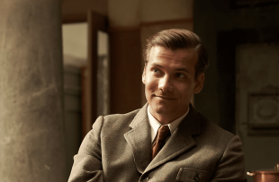 A man in a brown suit, sitting, smiles wryly at something we can't see in a publicity still for the show Hotel Portofino, on Binge.