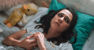 A young woman lies in bed with a concerned, confused look on her face, next to a very worn-looking duck teddy, in a publicity still for Such Brave Girls on Stan.