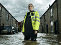 A woman police officer stands knee-deep in water on a flooded street in a publicity still for the new show After The Flood on BritBox.