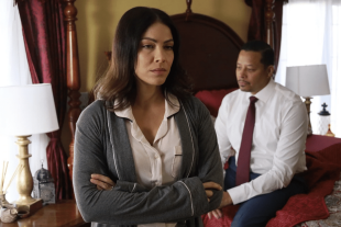 A woman stands in a bedroom with her arms crossed while a man in a business shirt and tie sits on the bed behind, in a publicity still for Skeletons in the Closet on AMC+ and Shudder.