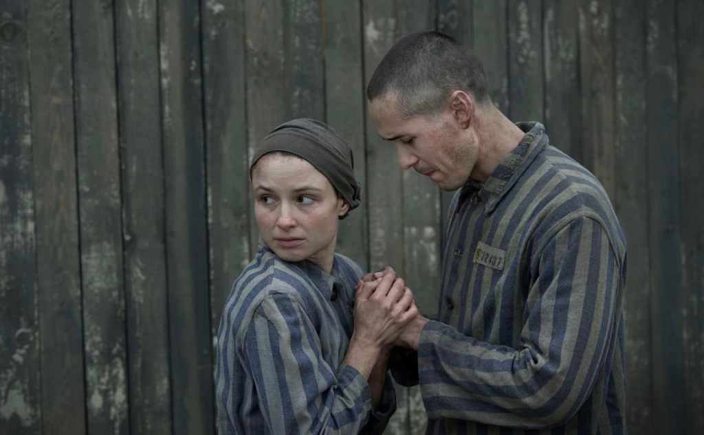 A woman and a man, both prisoners, hold hands, wearing concentration camp uniforms, in a still from The Tattooist of Auschwitz on Stan.