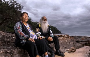 A First Nations woman and man sit on a rock on a beach facing the camera in a publicity still for Revealed: How to Poison a Planet on Stan.