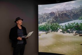 A older man wearing a black hat and black jacket holding a laptop and looking at a concave screen depicting a landscape with green shrubs.