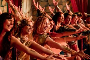 Pizazz perform 'Welcome to Troy' in the final show in HBO Max's 'Happiness'. A chorus line of smiling musical theatre performers smile at the audience, their arms outstretched behind and before them.