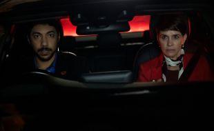 L-R: Hunter Page-Lochard and Tasma Walton in 'Reckless'. The photo depicts two First Nations actors, one male with a closely trimmed beard, the other female and wearing a red jacket with a black and white scarf tied around her neck. They are sitting side by side in the front seat of a car at night, with the male actor behind the steering wheel. He looks deeply worried, she looks intently into the rear view mirror, as if she is thinking furiously.
