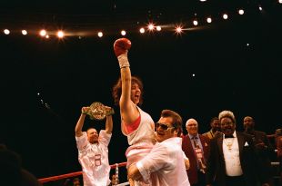 A scene from boxing biopic Christy showing Sydney Sweeney as the title character, gloved fist triumphantly raised as a man hoists her aloft in the boxing ring. Other men look on.