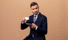 The Weekly's host, Charlie Pickering. A middle aged, fair-skinned man wearing a suit and tie, raises a tea cup from its saucer and looms glumly and sardonically at the camera.