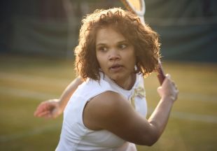 Lila McGuire as Evonne Goolagong in the ABC TV series Goolagong. A young Aboriginal woman in tennis whites is pictured mid-action on a tennis court, her bat raised.